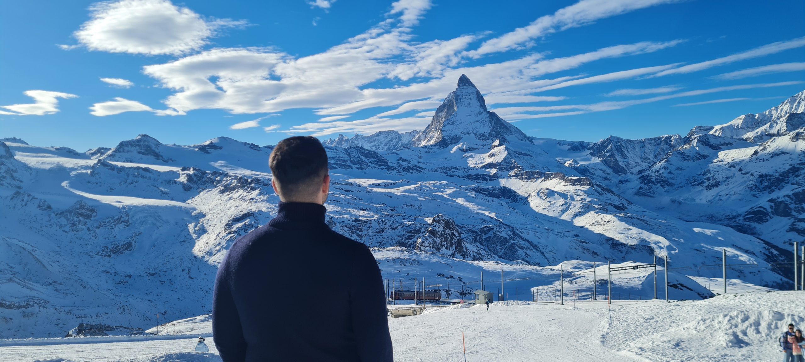A person in a dark jacket stands with their back to the camera, looking at the snow-covered Matterhorn mountain under a blue sky with scattered clouds.