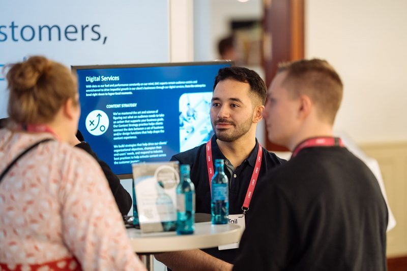 A person talking to two others at a professional event, with a screen in the background displaying information about digital services. The text to the left details the educational background of an individual with a Bachelor of Science in Applied Information Science from TH Köln, highlighting skills in SQL, SPSS, JavaScript, R, HTML, CSS, and experience as a freelance app developer.